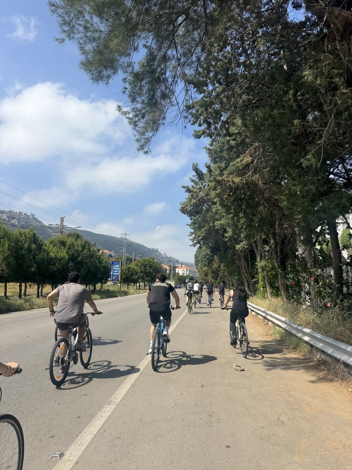 Bike Club Tripoli riders cycling down a tree-lined road past a guardrail, with an orange-roofed hillside village in the distance