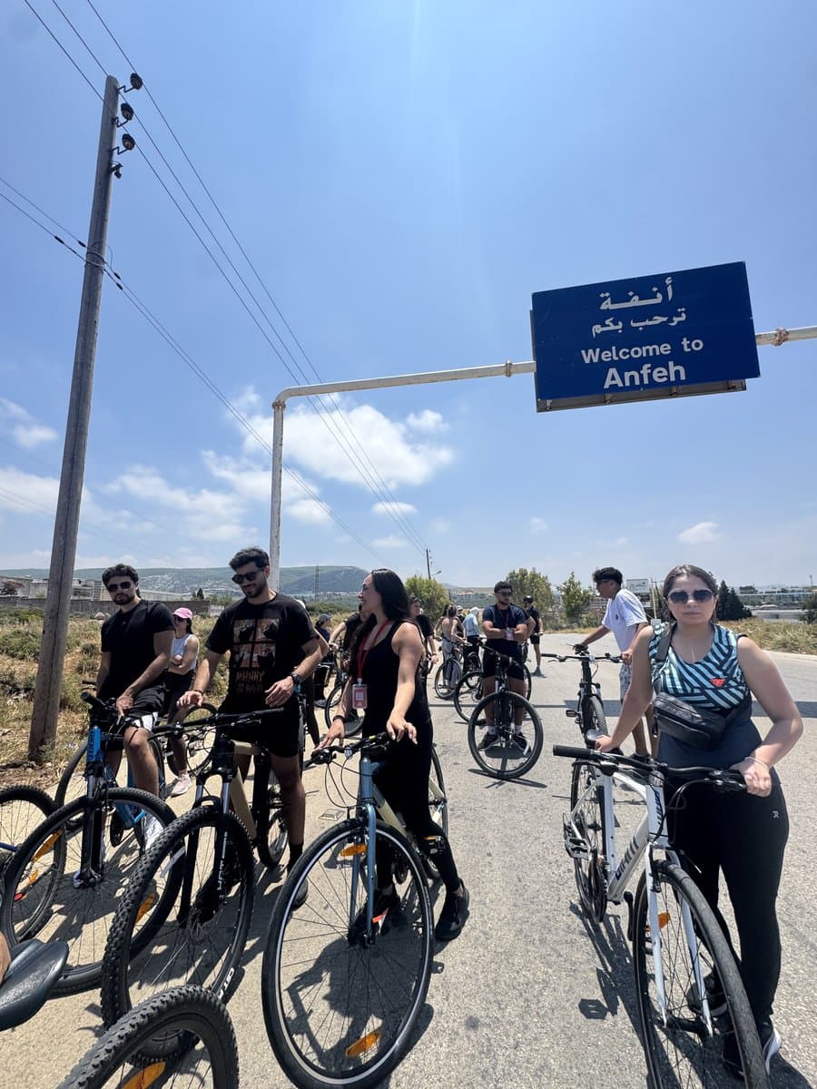 A group of Bike Club Tripoli riders pausing with their bikes beneath a blue road sign reading 'Welcome to Anfeh'