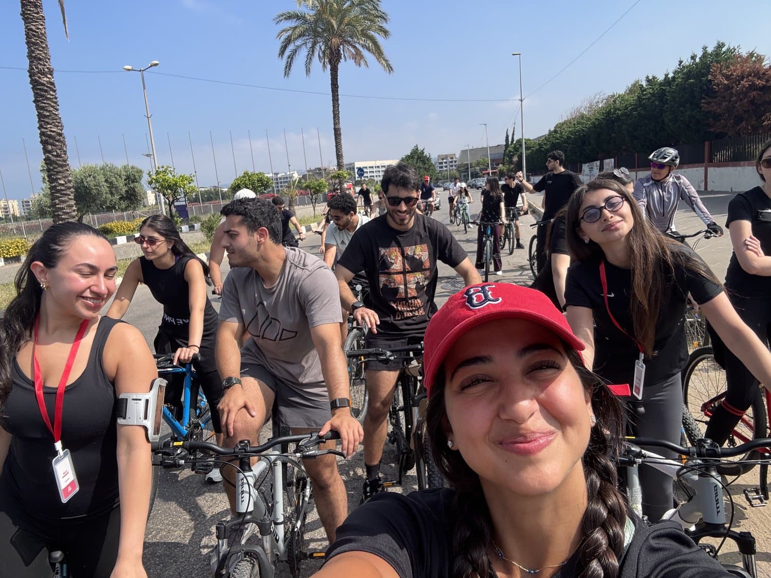 A group of Bike Club Tripoli riders smiling for a selfie on their bikes on a palm-lined coastal road, with the Tripoli coastline behind them