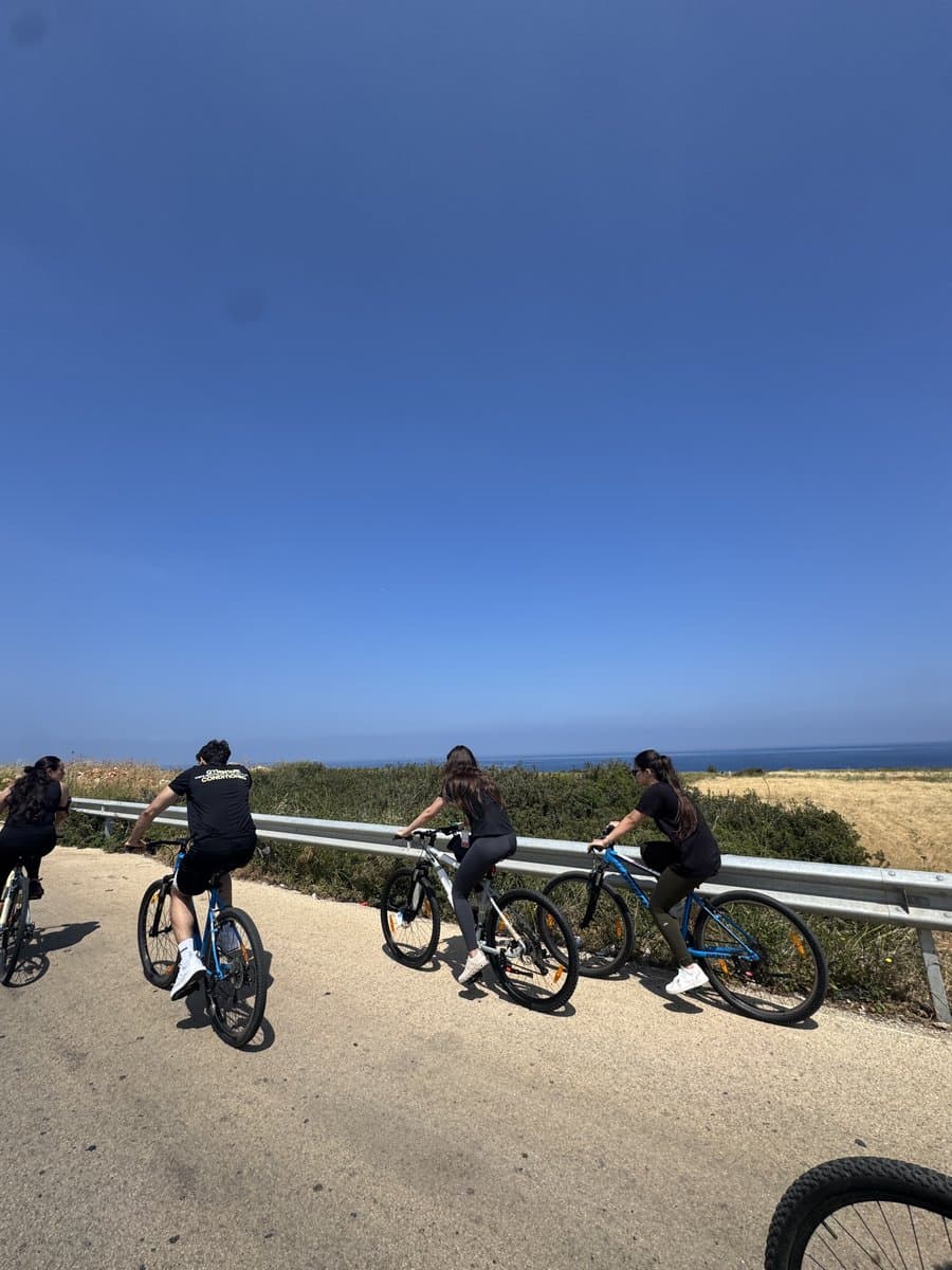 Three Bike Club Tripoli riders paused at a coastal guardrail overlooking the sea, with farmland on the other side of the road