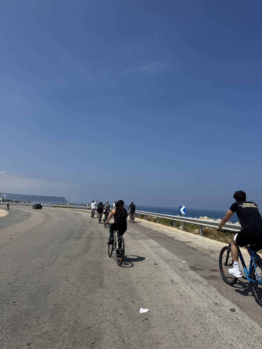 Bike Club Tripoli riders cycling along a coastal road beside the sea, with a headland visible ahead