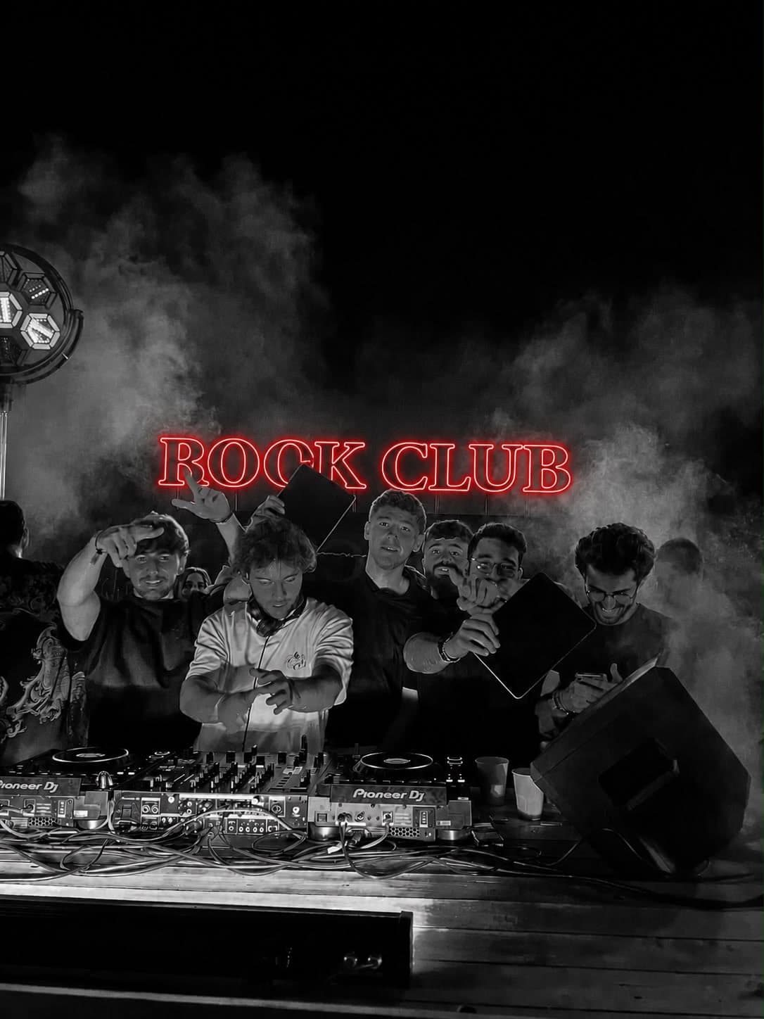 A group of six people posing at the DJ booth under a neon sign reading 'Rock Club,' with stage smoke effects, shot in black and white