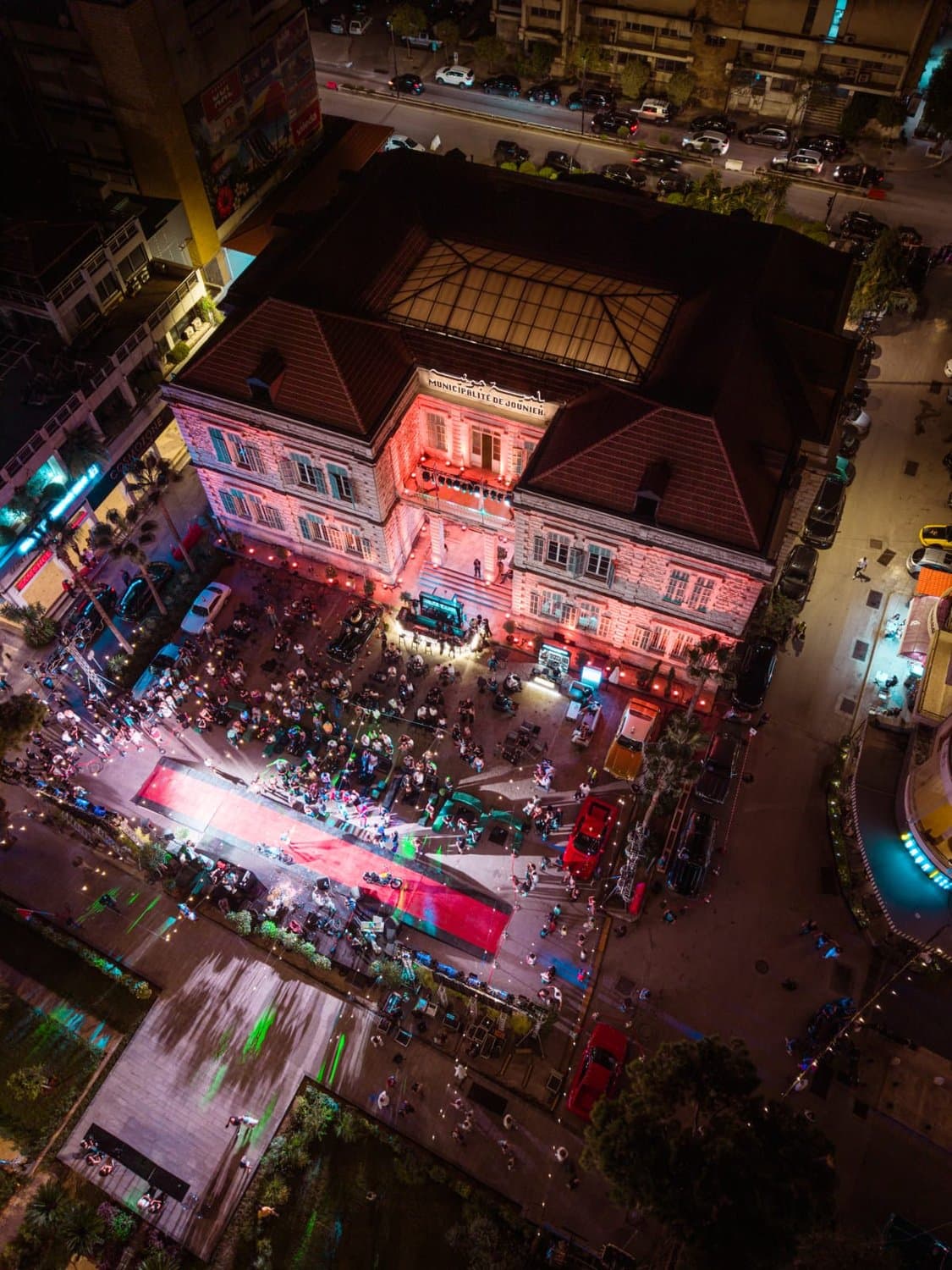 An aerial night view of Defile Classique's venue, a grand red-lit heritage building with a courtyard packed with attendees