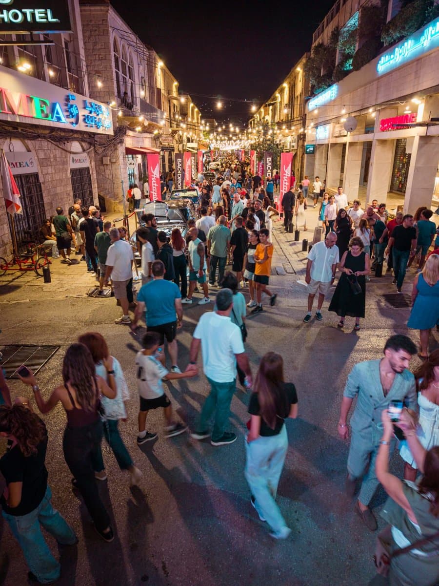 A crowded pedestrian street at night lined with classic cars and lit storefronts during Defile Classique