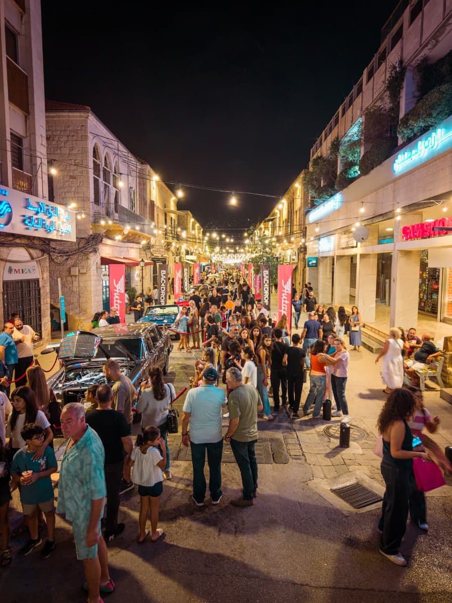 Classic cars parked along a festive, lantern-lit street as crowds walk past during Defile Classique