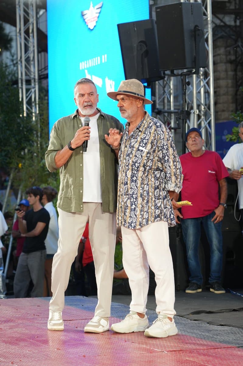 Two men on stage at Defile Classique, one speaking into a microphone, with a screen behind them reading 'Organized by'