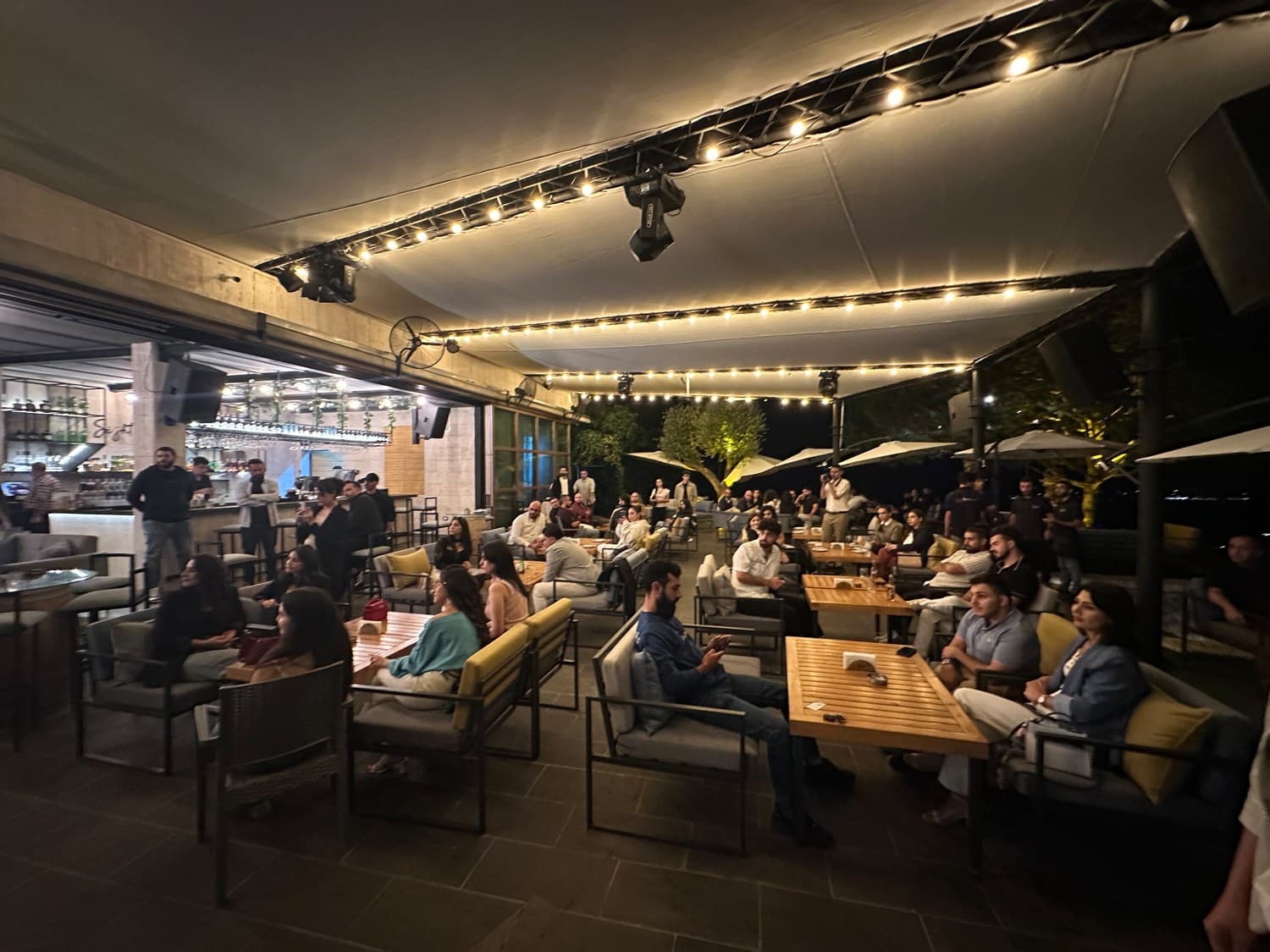 A crowded rooftop lounge terrace at night strung with warm string lights, packed with guests networking at Network With Jean in the Chouf