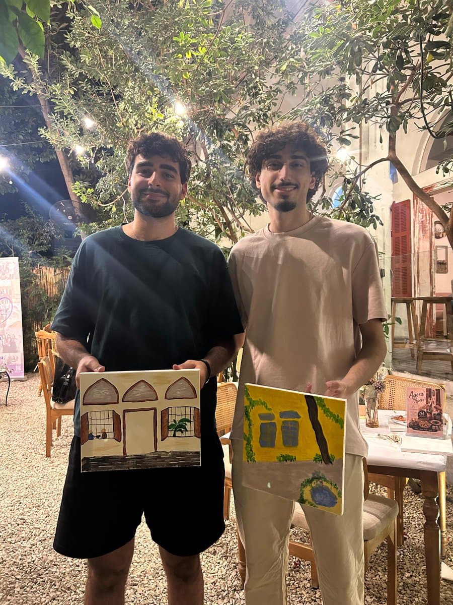 Two men at an evening Paint Club session under string lights, each holding up a finished painting