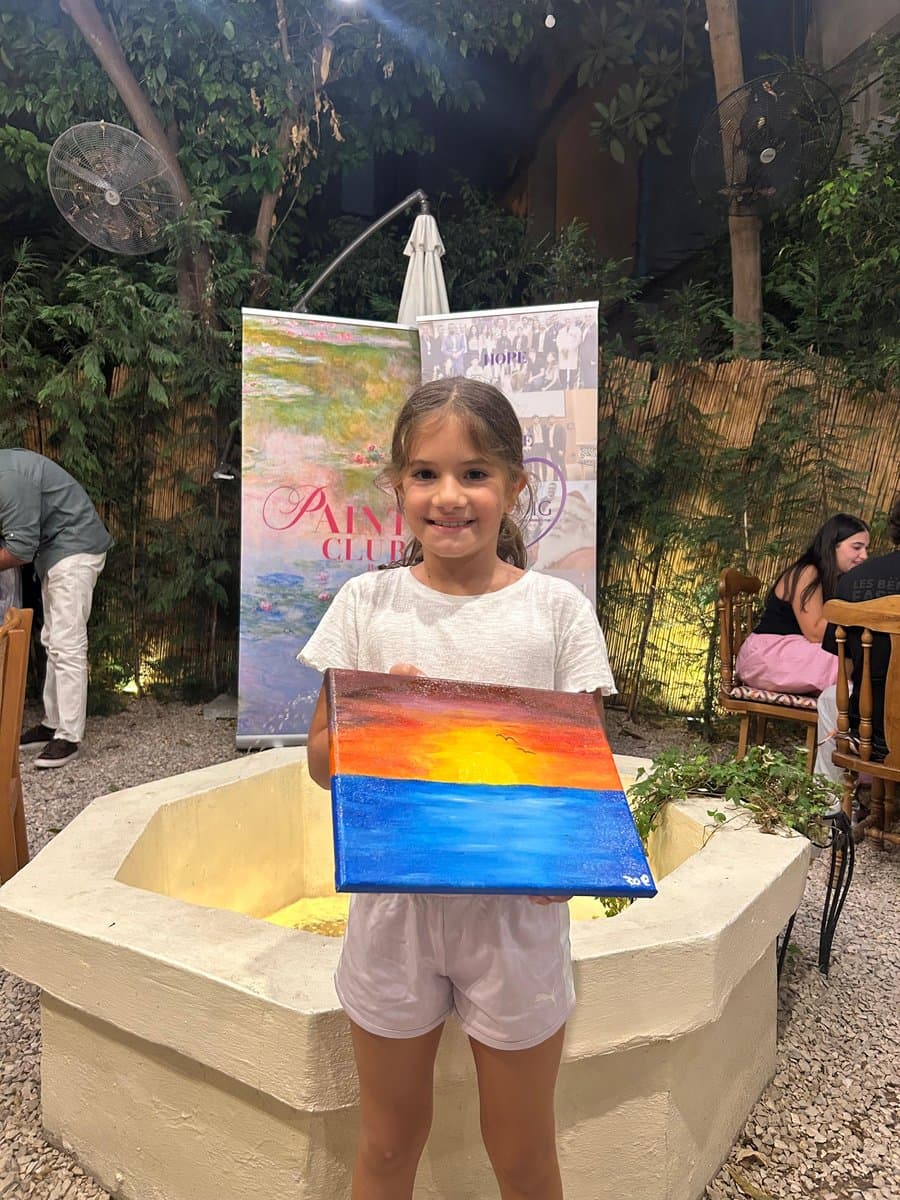 A young girl smiling while holding up a finished sunset painting at an evening Paint Club session, with a Paint Club banner visible in the background