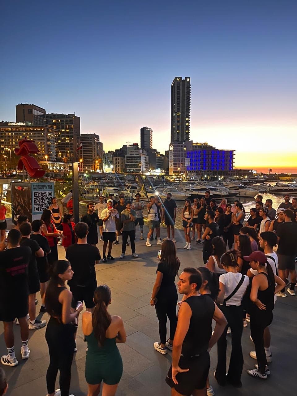 A crowd of Run Club Beirut runners gathered at dusk along Beirut's waterfront, with the city skyline and a marina behind them