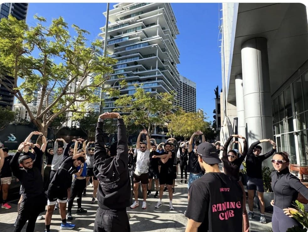 A large group of Run Club Beirut runners doing a pre-run stretch with their arms raised overhead, on a sunny street lined with trees and modern high-rise buildings, with one runner's black t-shirt printed with the club's name in pink lettering