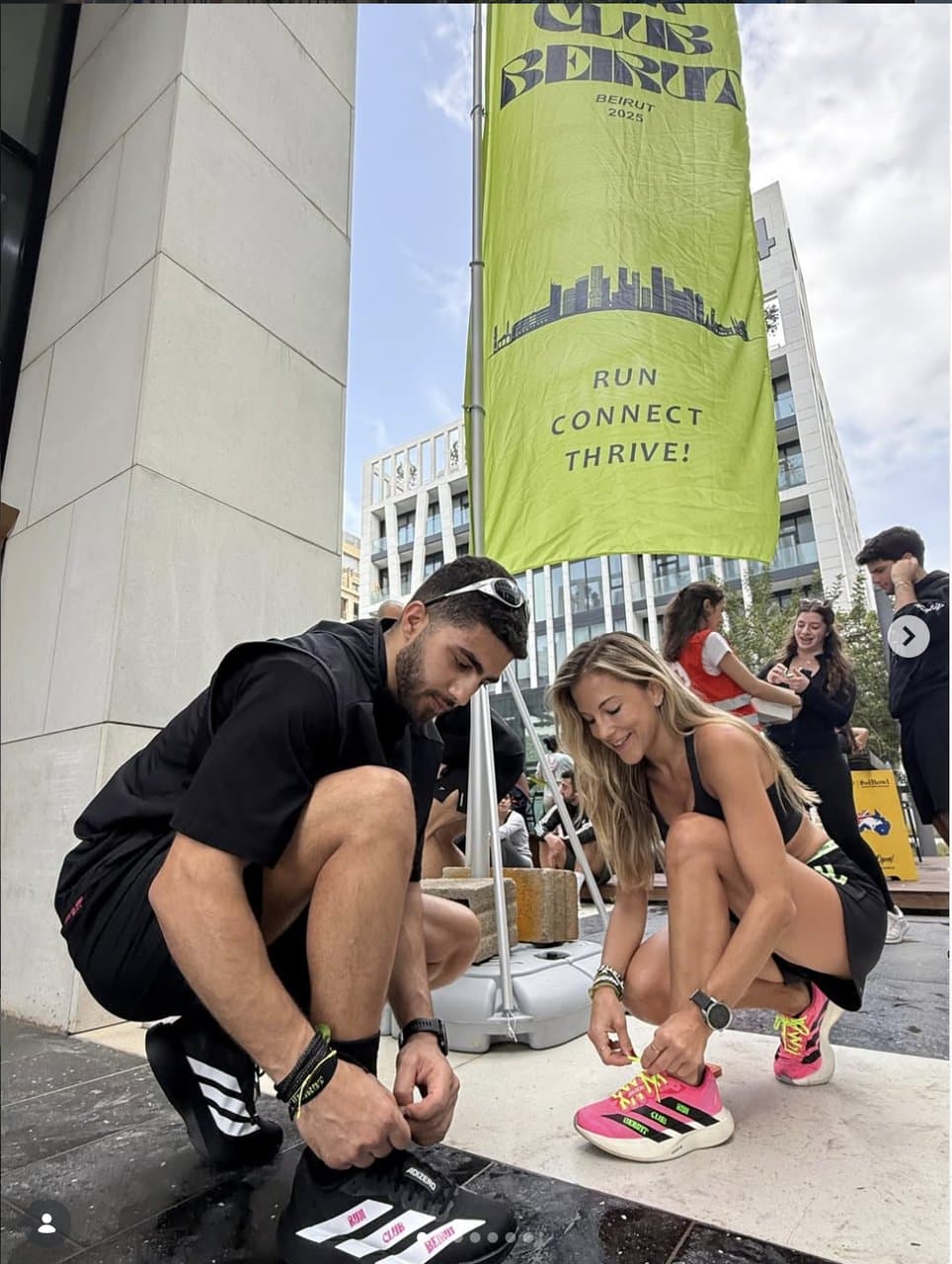Two runners tying their shoes beneath a Run Club Beirut flag reading 'Run, Connect, Thrive', with the Beirut skyline behind them