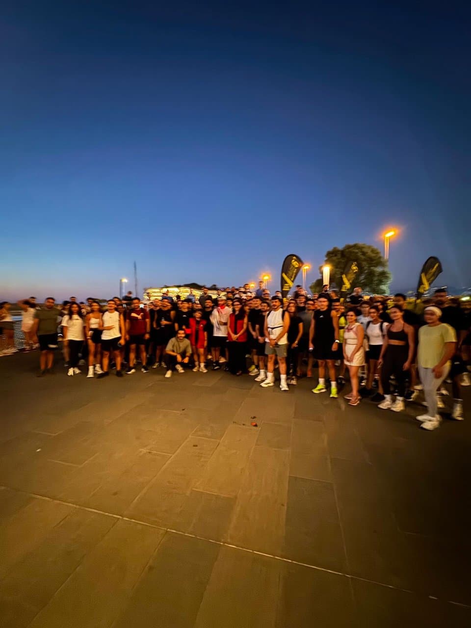 A large crowd of Run Club Beirut runners standing together at dusk on a waterfront promenade, with branded flags and string lights behind them
