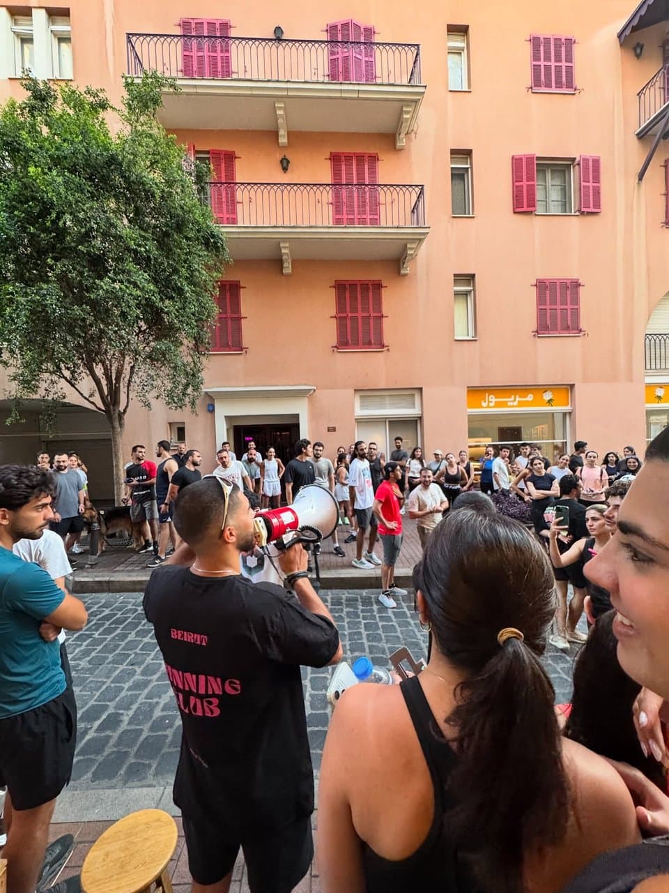 A Run Club Beirut leader briefing a crowd of runners through a megaphone on a street in front of a pink building with red shutters
