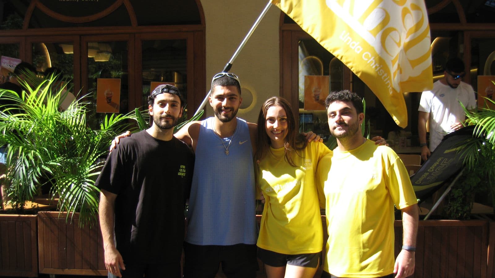 Four organizers posing together with the yellow Sabah El Kheir flag, printed with Linda Christina's name, outside the café