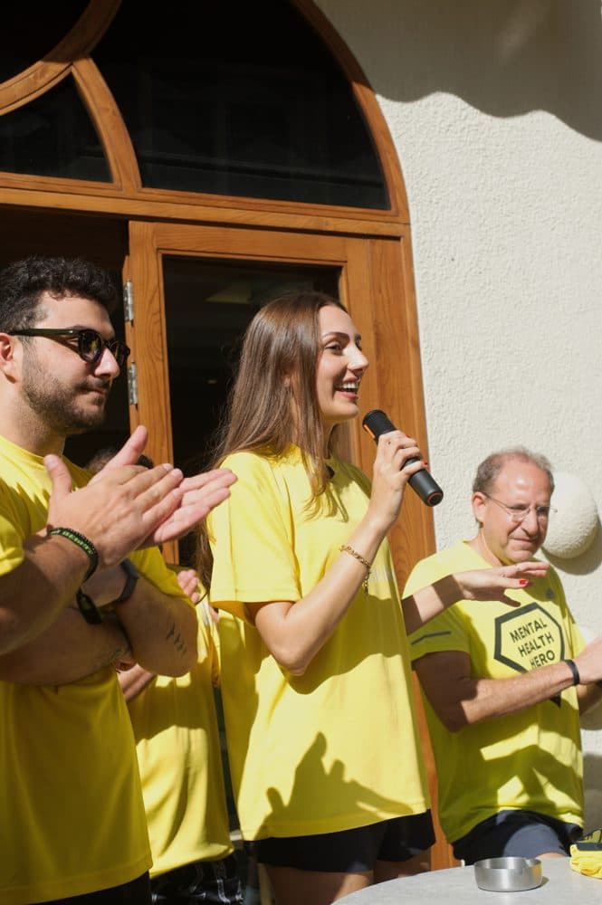 Linda Christina speaking into a microphone in front of the café, with attendees in yellow Sabah El Kheir Run t-shirts applauding beside her