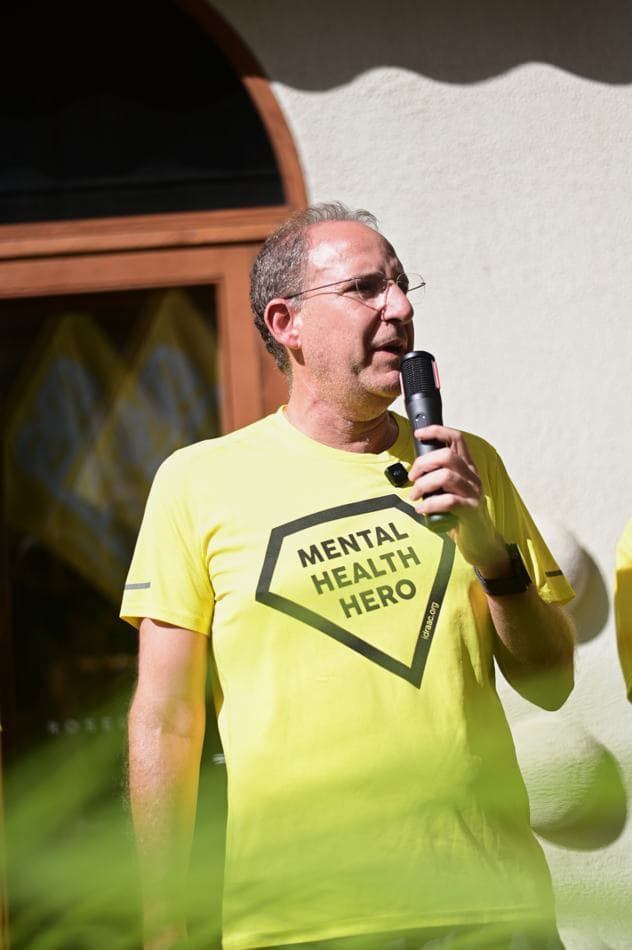 A speaker in a yellow "Mental Health Hero" t-shirt addressing the group with a microphone before the run, Roselane Café signage visible behind him