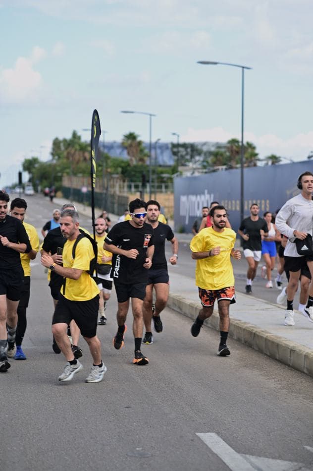 A larger group of runners in yellow and black t-shirts running along a wide street with a branded flag overhead
