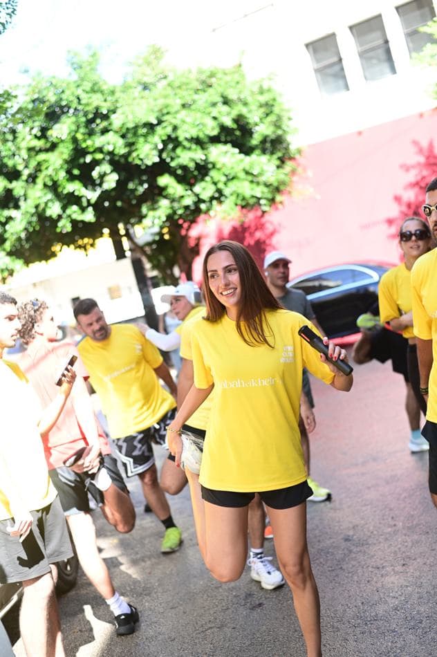 A runner in a yellow Sabah El Kheir Run t-shirt smiling while running past parked cars and trees
