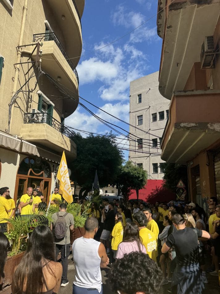A large crowd wearing yellow Sabah El Kheir Run t-shirts gathered in a narrow Beirut street lined with balconies, with a speaker holding a microphone