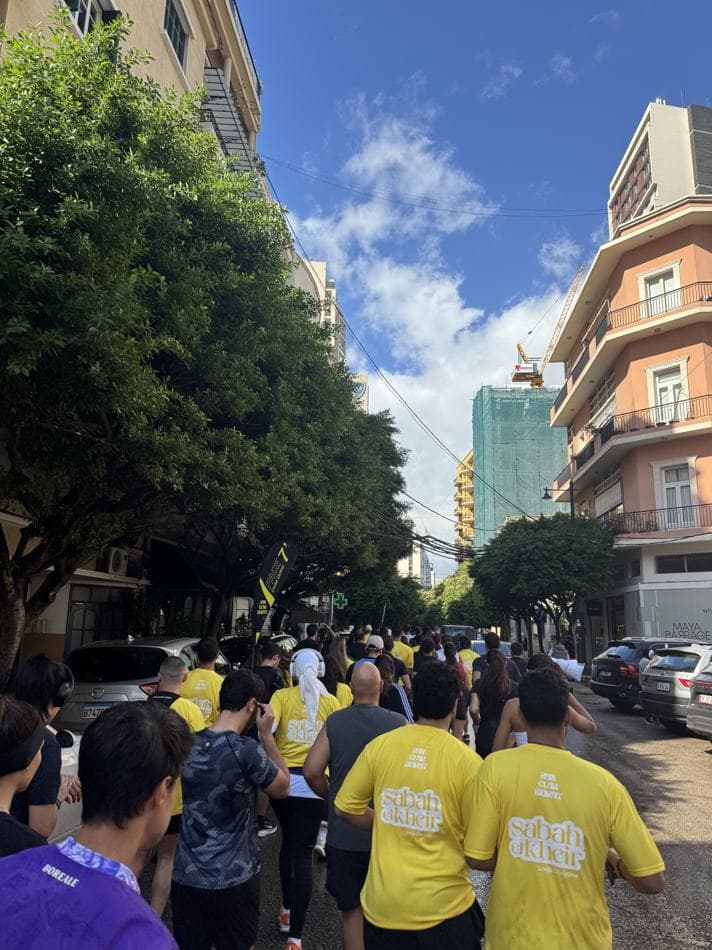 Runners and attendees in yellow t-shirts walking down a Beirut street with a branded flag visible above the crowd