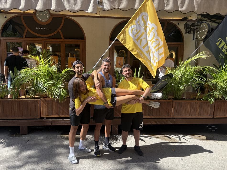 A group of attendees posing playfully together, one person being lifted, with the Sabah El Kheir flag in the background outside a café