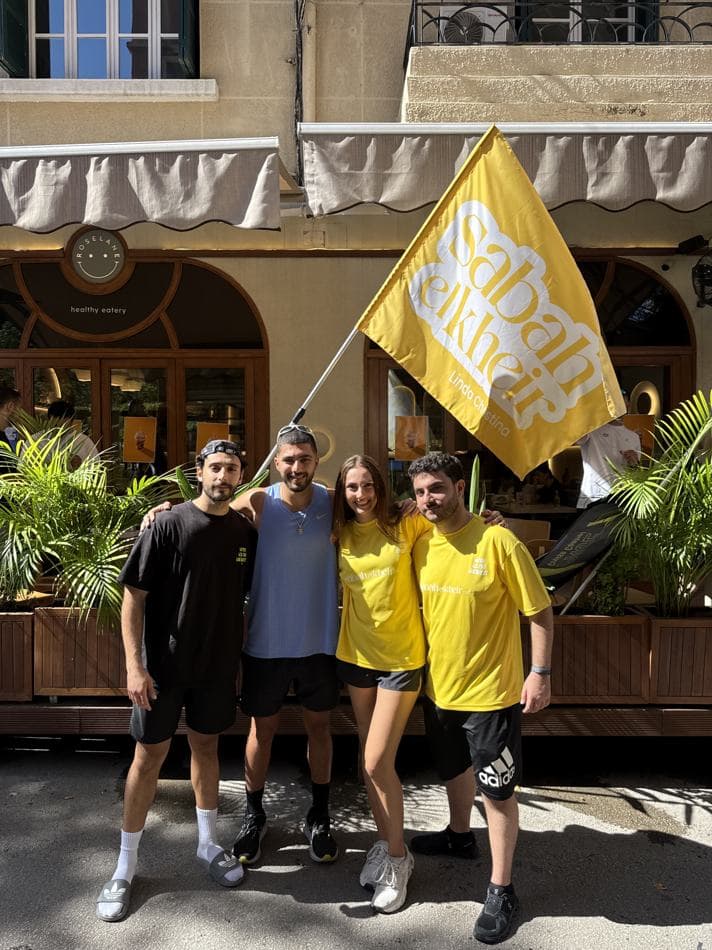 Four attendees posing with the yellow Sabah El Kheir flag, printed with Linda Christina's name, outside the café