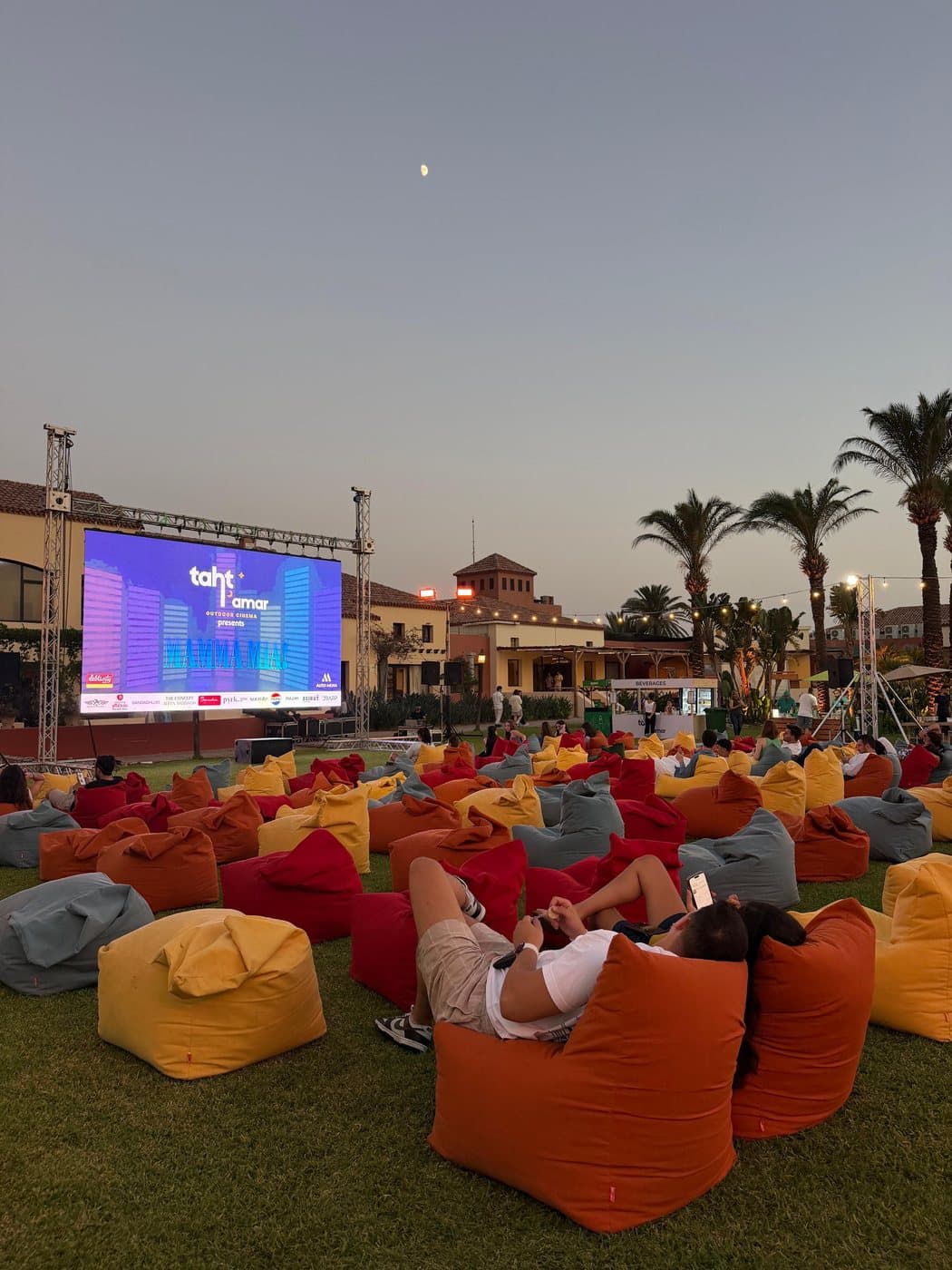 Taht El Amar's outdoor cinema screen lit up at dusk, with a couple watching from their beanbags in the foreground