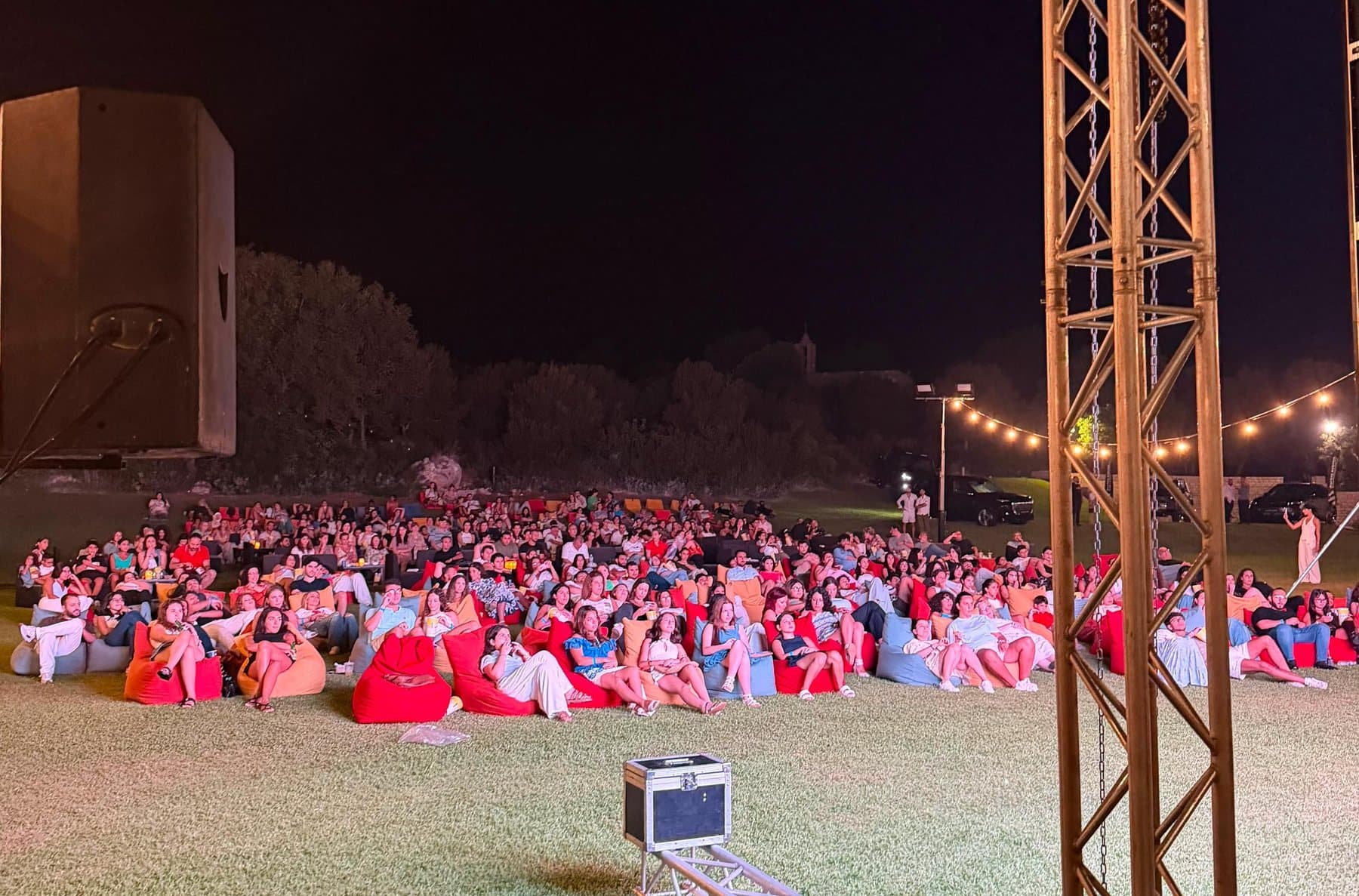A full crowd seated across beanbags at night, under the lighting truss, at a Taht El Amar screening