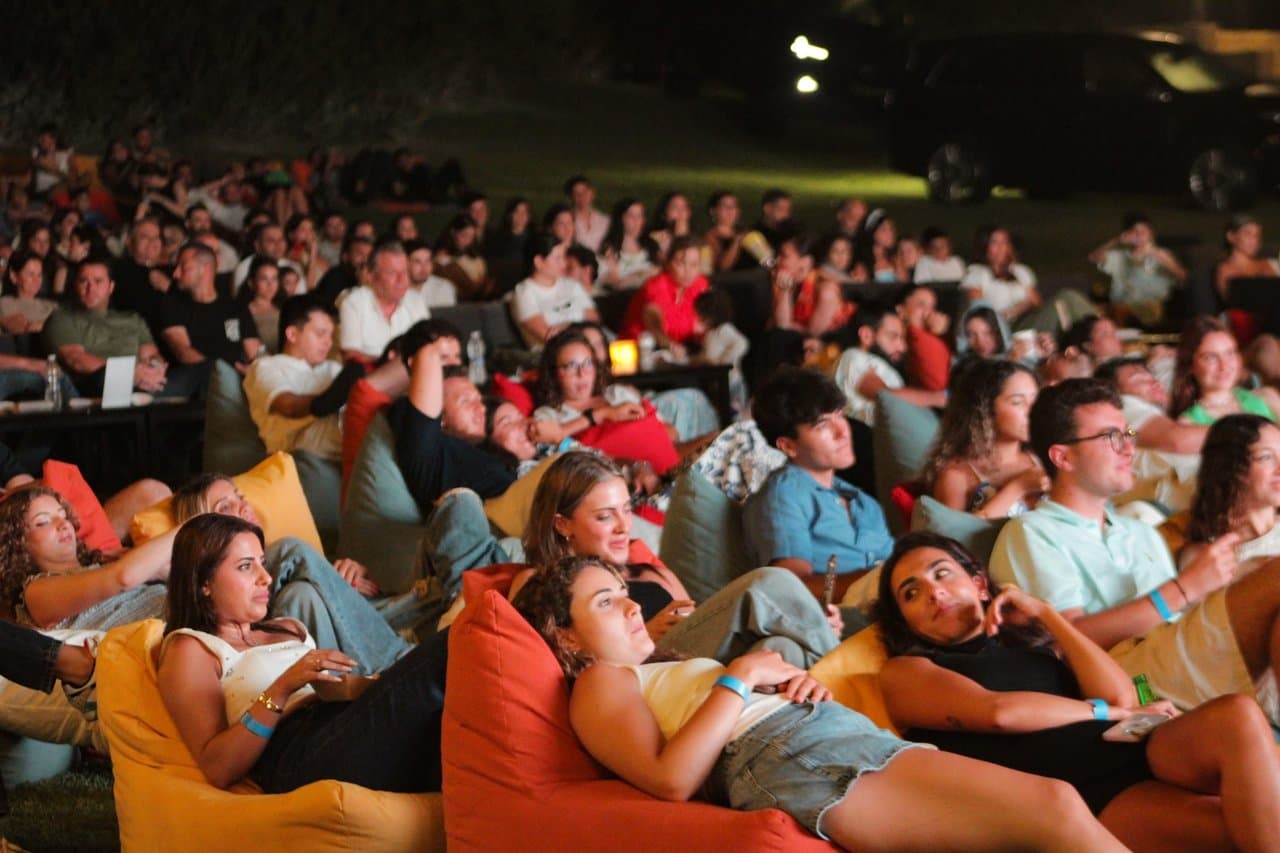 A large crowd seated across beanbags at night, with car headlights and taillights visible on the road behind the venue