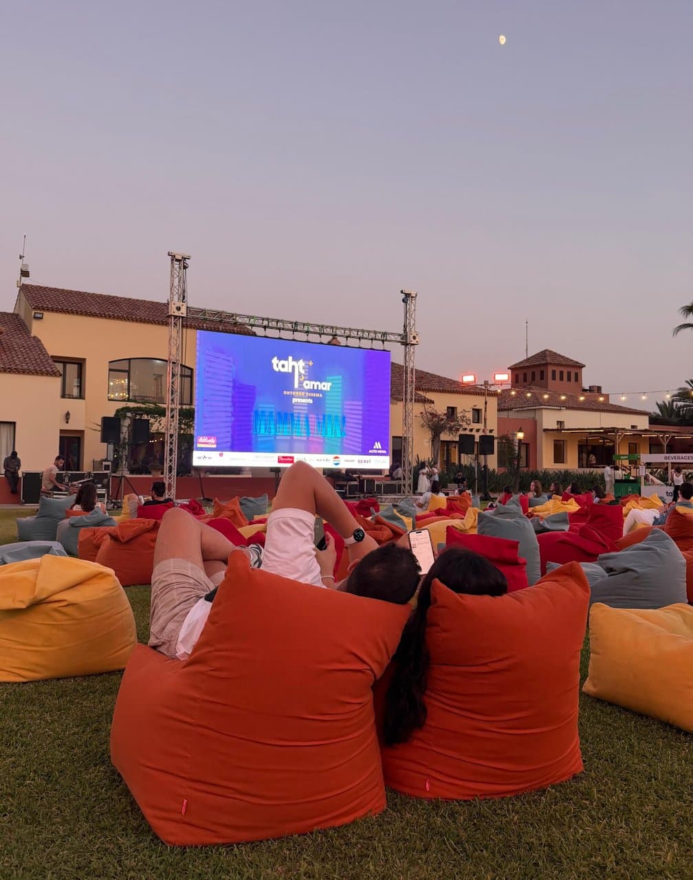 A wide dusk view of the Taht El Amar screen showing Grown Ups, with a village-style building, garden and the moon in the background