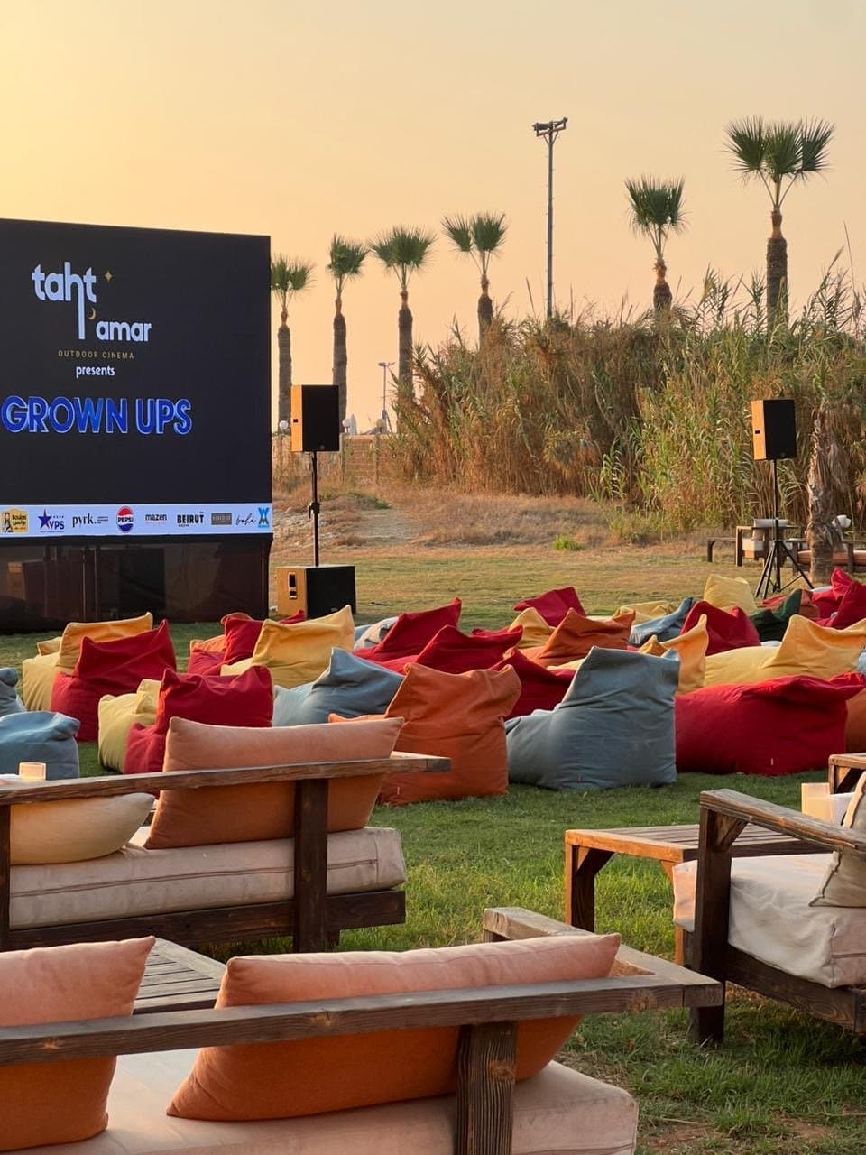 Empty VIP lounge sofas and rows of beanbags facing the Taht El Amar screen showing Grown Ups, with palm trees behind, before guests are seated